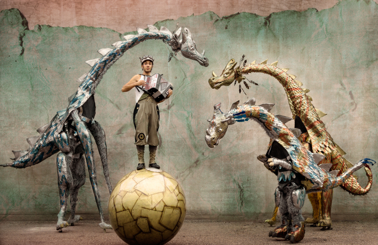 hombre con acordeón sobre una bola de gran tamaño, tres figuras de dragones a su alrededor